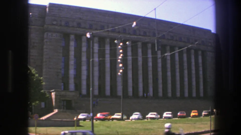 1975 Sweden 8mm view of the municipal hall facade, with vertical columns, parked 1970s cars, overhead wires, lawn, and street foreground.