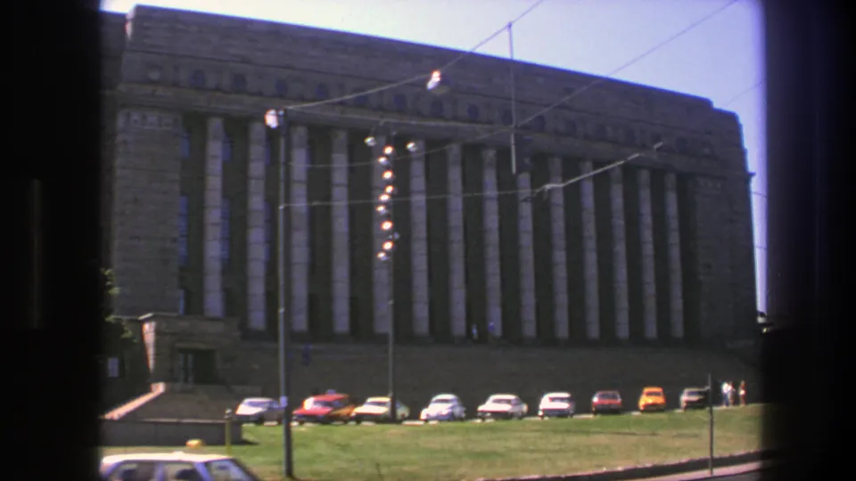 1975 Sweden frame showing the stone public hall, tall columns, parked cars, trolley wires, lawn, and a passing car at the bottom edge.