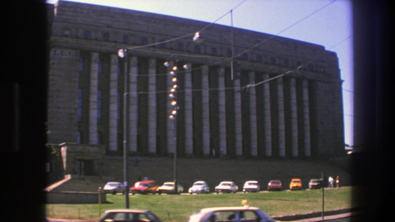 Large stone building with vertical columns, grass foreground, parked cars, and overhead wires.