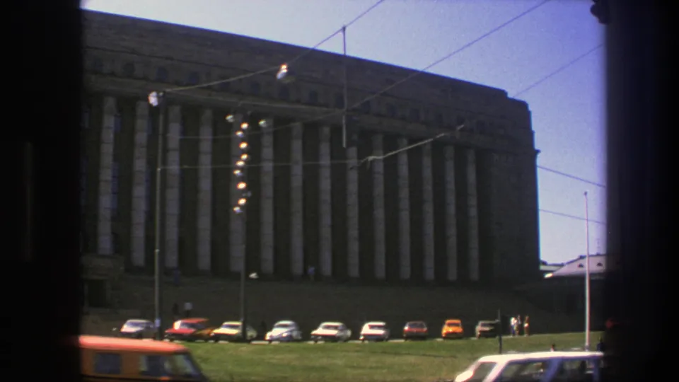 1975 Sweden 8mm frame showing the columned building from a moving street view, with parked cars, wires, poles, and partial passing traffic.