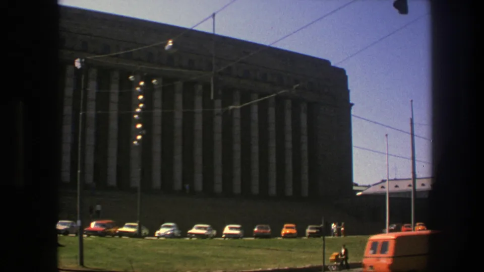 Late clip frame of the 1975 Sweden building exterior, with columns in shadow, parked cars, overhead wires, and an orange vehicle entering at right.
