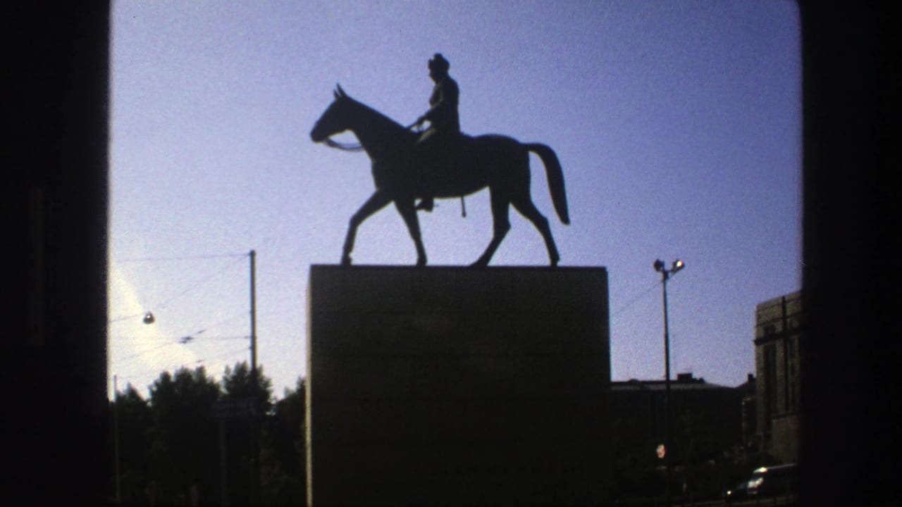 Backlit 8mm view of a Swedish city equestrian monument, framed with tram wires, a lamppost, and dark building forms.
