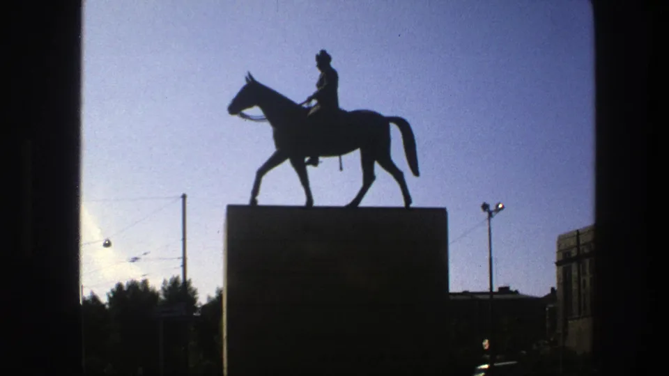 Closer 1975 Sweden home-movie frame of the equestrian statue, with the pedestal, overhead wires, and street lighting visible.