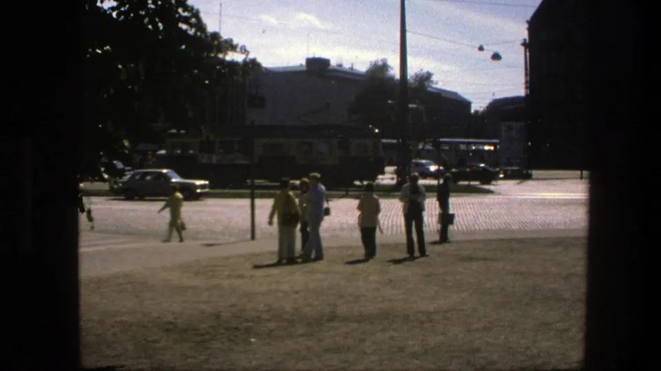 Wider 1975 Sweden street view showing pedestrians in a public square, period vehicles, overhead wires, and surrounding buildings.