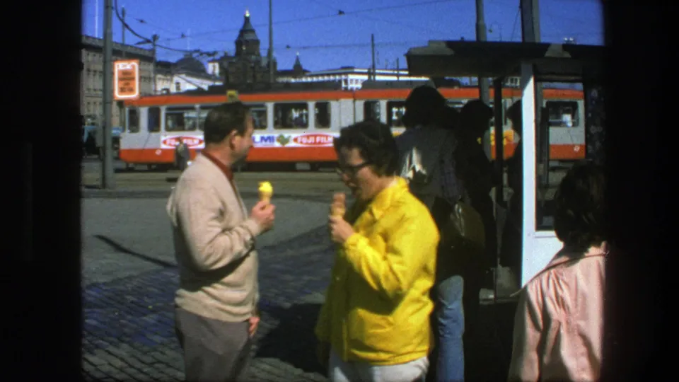 1975 Sweden home-movie frame showing adults with ice cream cones near a tram stop as a red-and-white tram passes behind them.