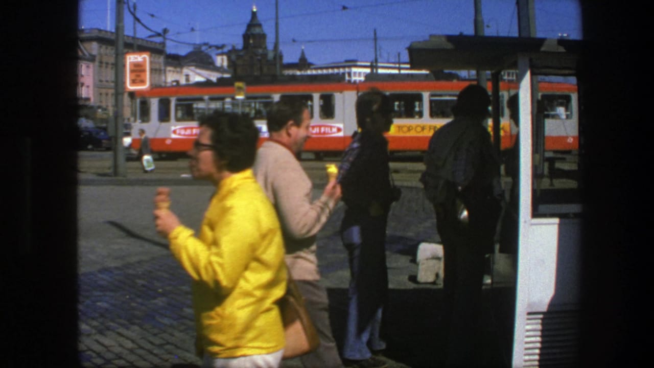 People stand near a tram stop as a red-and-white tram passes in the street.