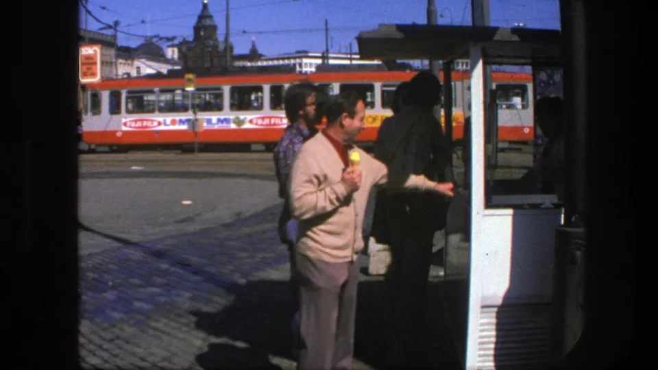 1975 Sweden frame showing a man with an ice cream cone, other pedestrians, a tram stop shelter, and a red-and-white tram behind them.