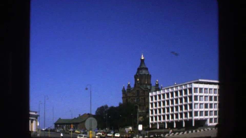 1975 Sweden street view showing a dark historic towered building beside a white modern building, with cars and street signs below.