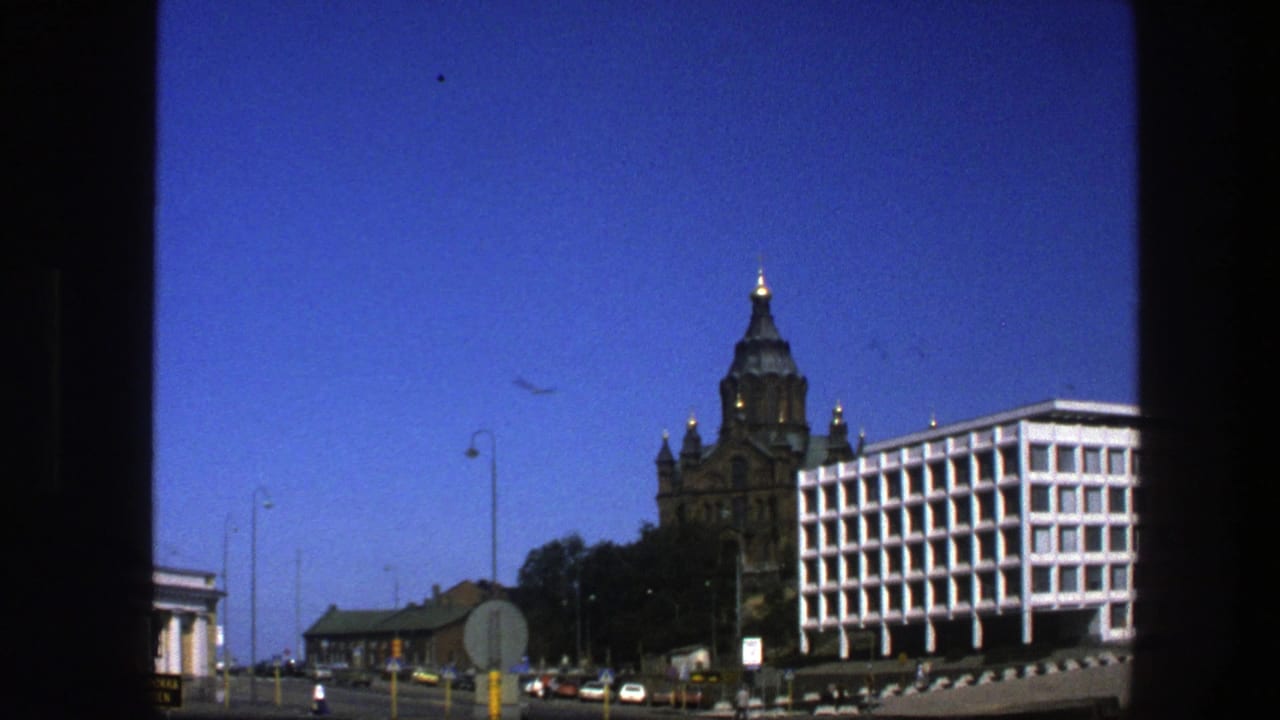 City street seen through a dark window frame with ornate tower, modern white building, cars, signs, and blue sky.