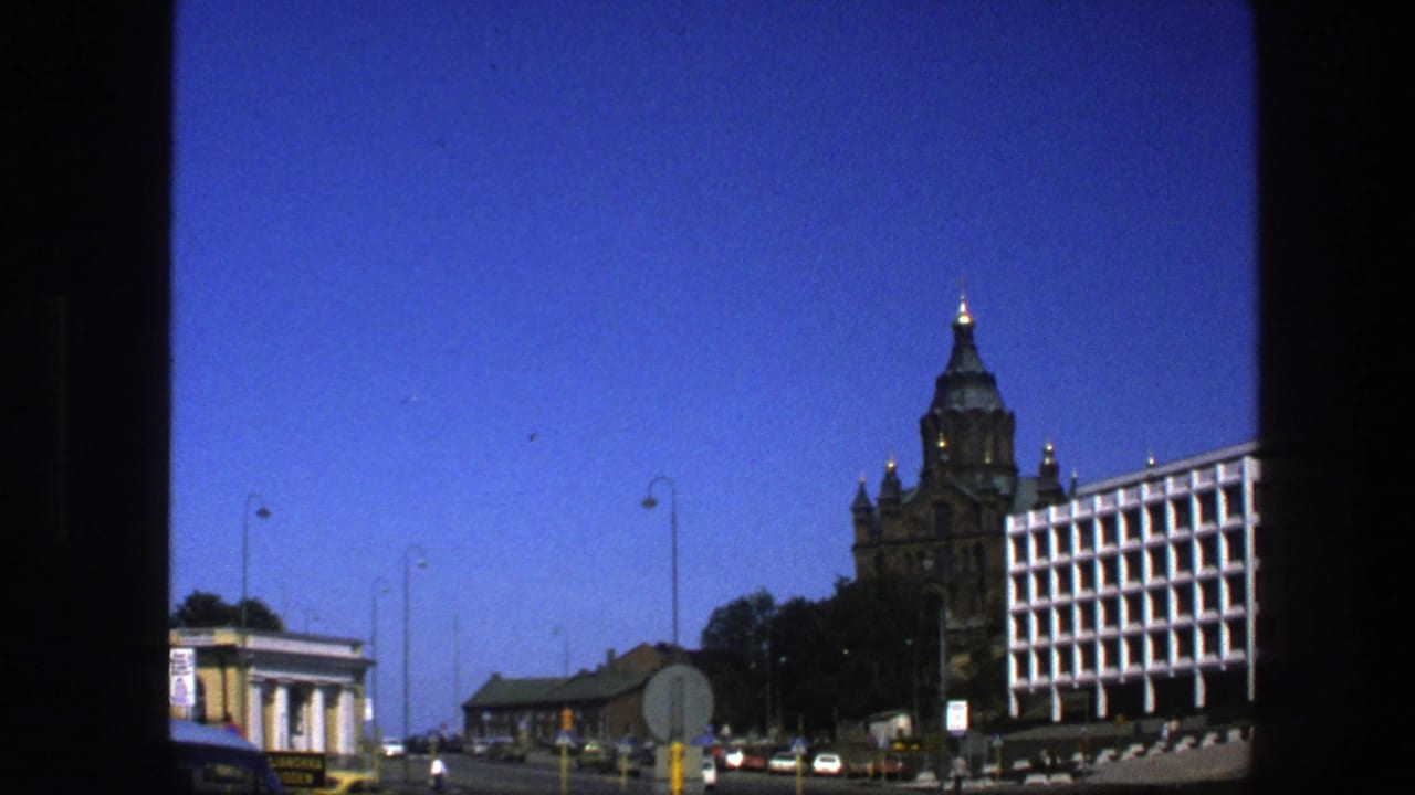 1975 Sweden street scene with a bird crossing the sky above an ornate tower, modern white building, road signs, cars, and lamp posts.