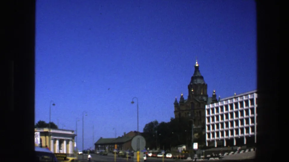 1975 Sweden street scene with a bird crossing the sky above an ornate tower, modern white building, road signs, cars, and lamp posts.