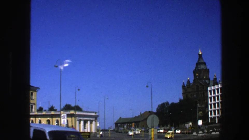 1975 Sweden city street with an ornate towered building, traffic signs, cars including a yellow taxi-like vehicle, lamp posts, and blue sky.