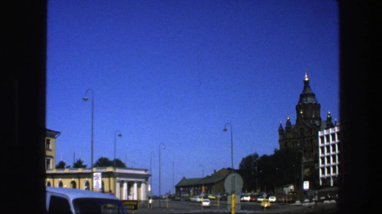 1975 Sweden 8mm frame showing a city street seen through a dark window frame, with cars, older buildings, and a distant tower.