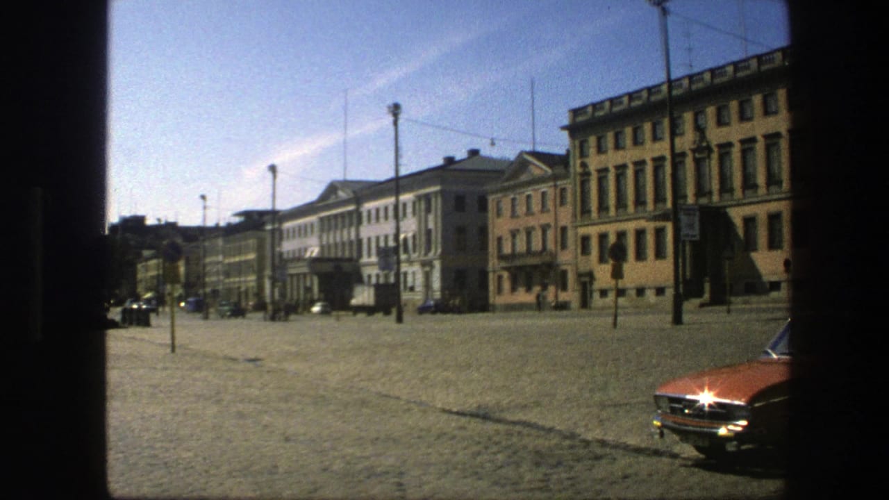 1975 Sweden 8mm frame of a sunlit cobblestone square with historic facades, overhead wires, streetlamps, and a red car partly visible.