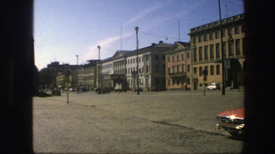 1975 Sweden 8mm frame showing a wide cobblestone plaza with rows of older buildings, utility poles, overhead wires, and a red car in front.