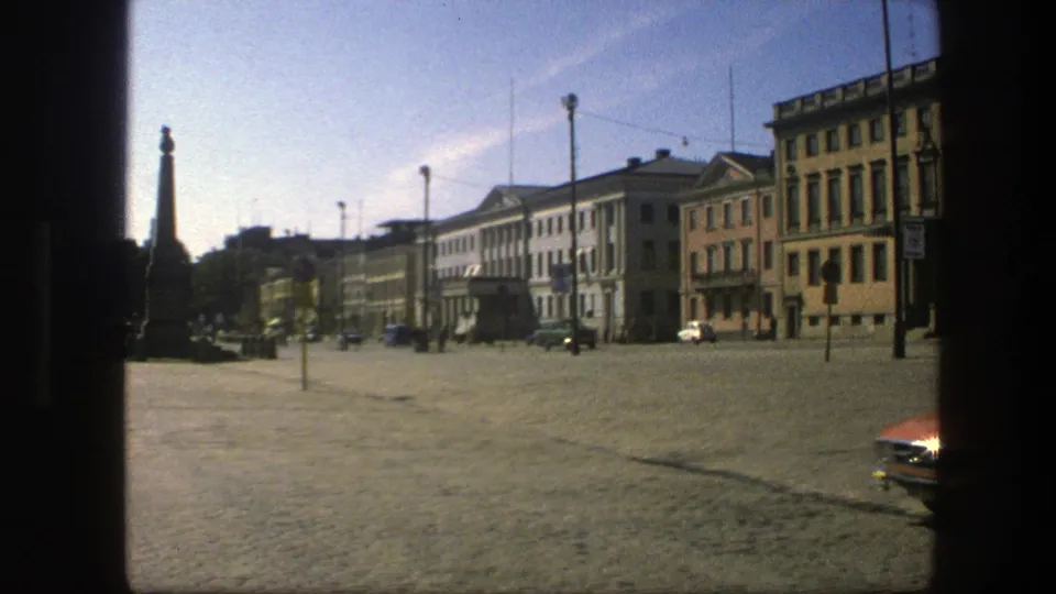1975 Sweden 8mm frame of a sunlit city square with older facades, overhead wires, a monument-like silhouette, and part of a red car.