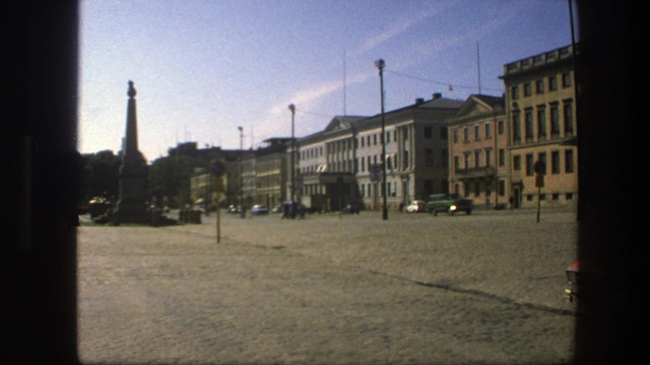 1975 Sweden street view of a cobblestone square with an obelisk, distant pedestrians, cars, and classical buildings under a clear sky.