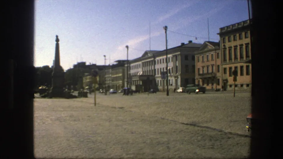 1975 Sweden street view of a cobblestone square with an obelisk, distant pedestrians, cars, and classical buildings under a clear sky.