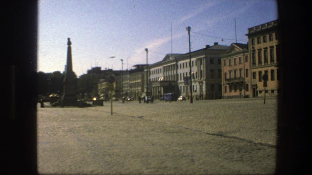 Wide 1975 Swedish urban square with an obelisk at left, small figures walking, period cars, and pale building facades.
