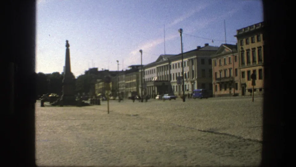 Swedish town square scene from 1975 with a blue van near the buildings, a monument at left, pedestrians, and soft 8mm color.
