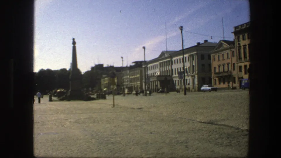 Late clip frame of a 1975 Swedish cobblestone square, showing an obelisk, a white car, distant pedestrians, and civic buildings.