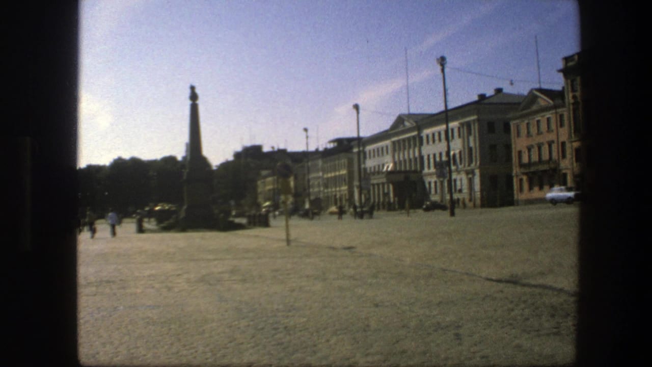 1975 Sweden home-movie frame showing a sunlit cobblestone square, tall stone monument, distant pedestrians, cars, and multi-story buildings.