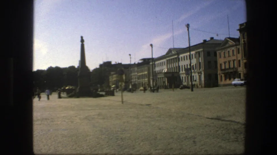 1975 Sweden home-movie frame showing a sunlit cobblestone square, tall stone monument, distant pedestrians, cars, and multi-story buildings.