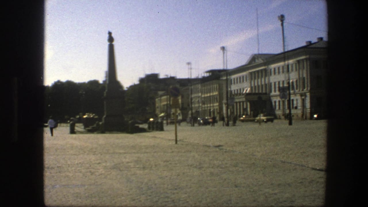 Public square with a tall monument at left, pedestrians, cars, and a row of buildings under clear sky.