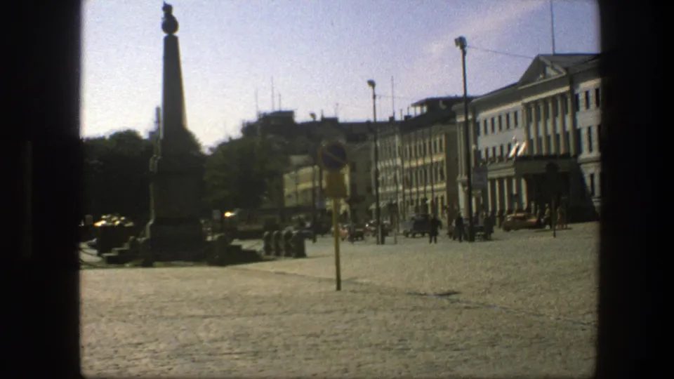 1975 Sweden home-movie frame zoomed toward a yellow street sign pole, with the monument, cobbled plaza, cars, and pedestrians behind it.