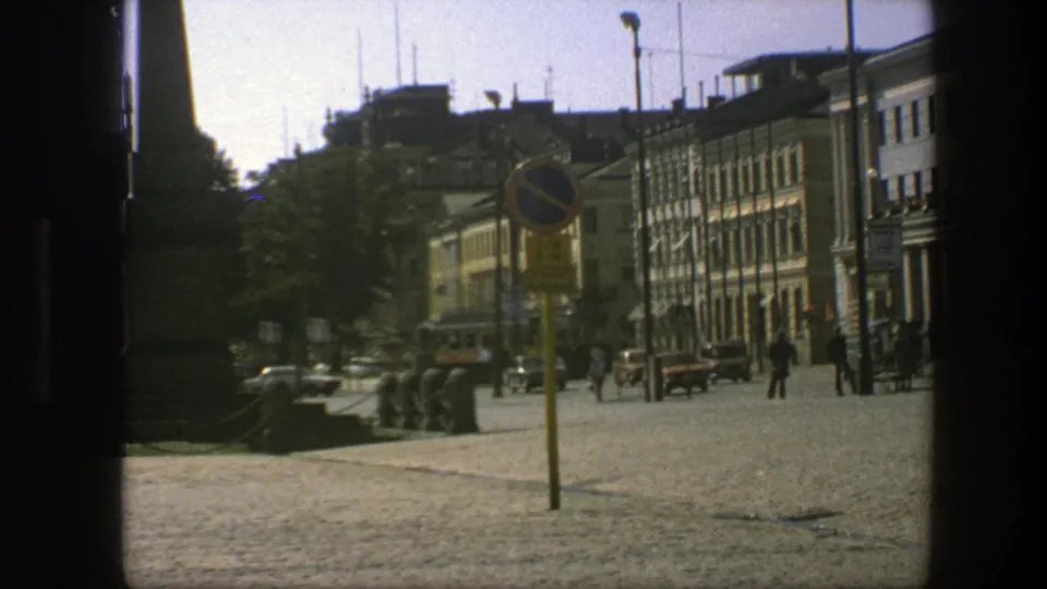 1975 Sweden frame showing the zoomed public square with a yellow sign pole in front, buildings at right, cars, pedestrians, and the monument partly cropped.