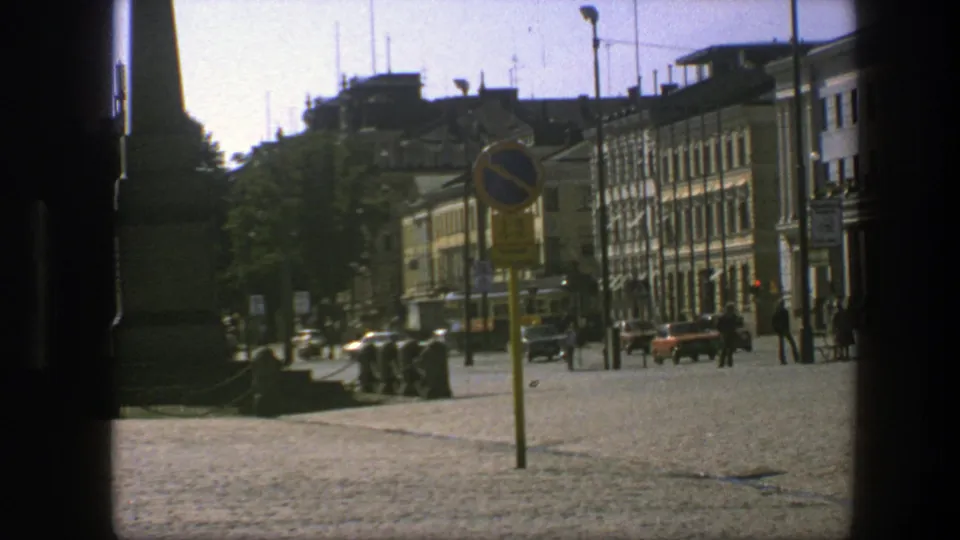 1975 Sweden street view with pedestrians crossing a cobblestone area near cars, buildings, and a prominent roadside sign.