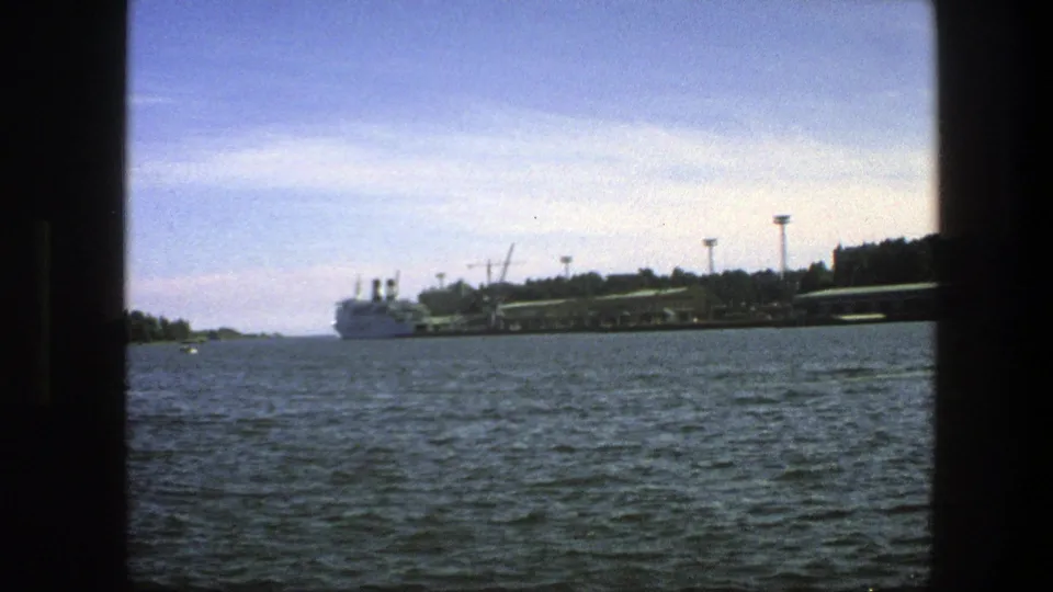 Wide 1975 Swedish harbor view showing a distant ferry, long pier, cranes, shoreline, and rippled water.