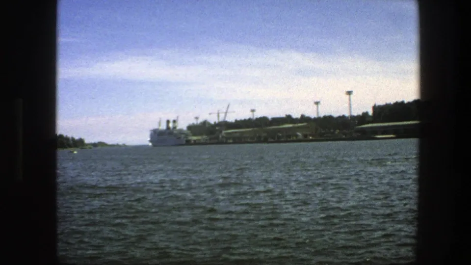 1975 home-movie frame of a Swedish waterfront, with choppy water, a white ship, trees, dock structures, and a bright sky.