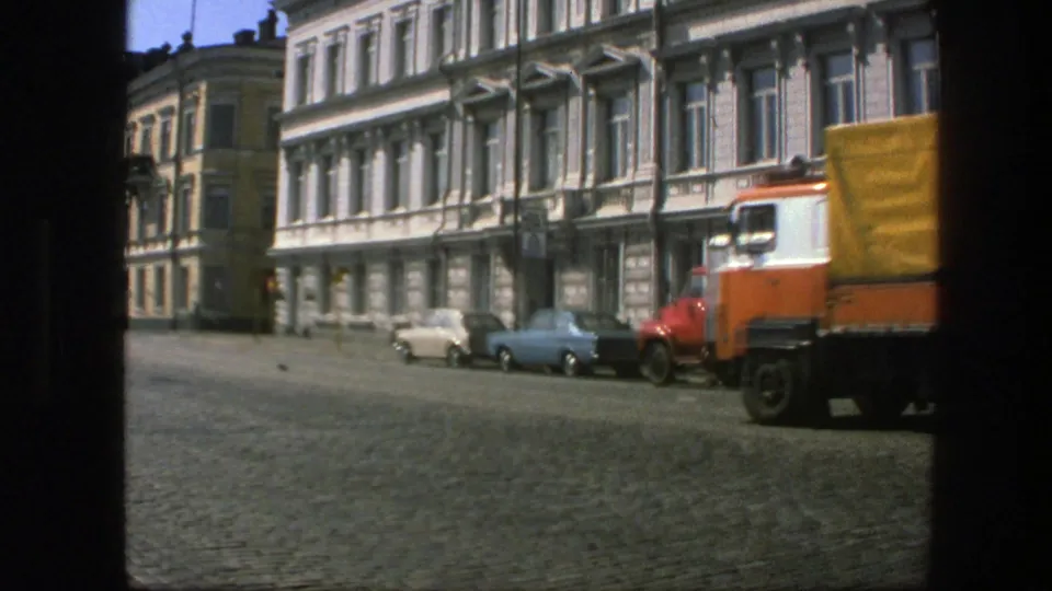 1975 Sweden 8mm frame of a covered goods lorry moving along a cobblestone street near older multi-story buildings.