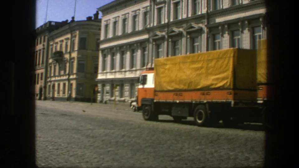 The yellow-covered lorry moves through the frame on a Swedish cobblestone street, with older facades and parked cars behind it.
