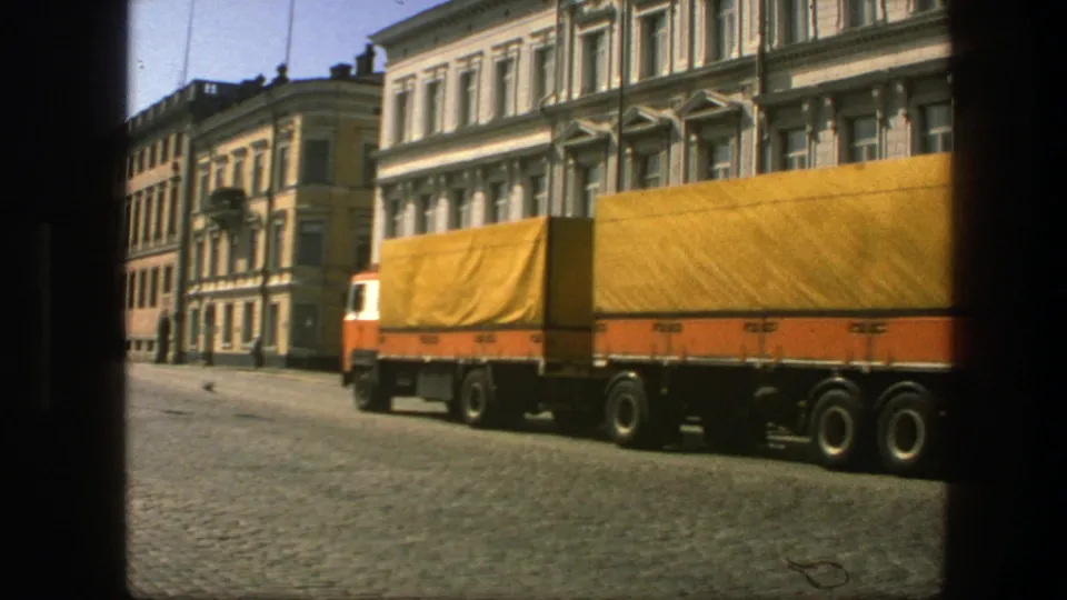 Side view of the covered goods lorry crossing the frame in 1975 Sweden, with cobblestones and older urban buildings visible.