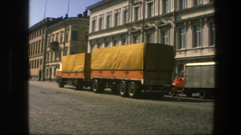 The lorry exits the street view near a red vehicle, with Swedish urban facades and cobblestones in the background.