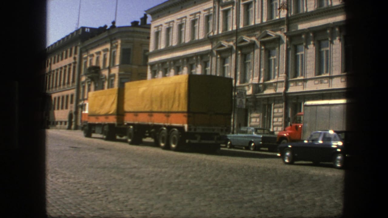1975 Sweden street view with a yellow-tarp freight truck, parked sedans, a red truck cab, and ornate white buildings seen through dark framing.