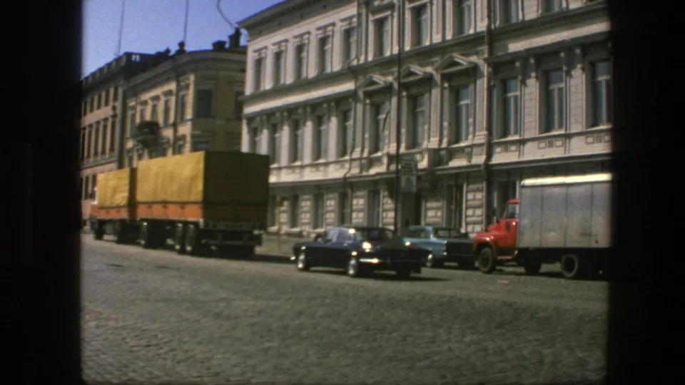 Yellow-tarp freight truck on a Swedish cobblestone street in 1975, with parked cars, a red truck cab, and ornate building facades in view.