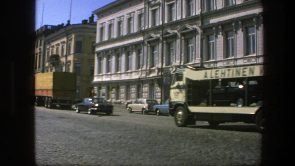 A tan tour bus with visible side lettering passes a yellow truck and parked cars on a 1975 Sweden cobblestone street.