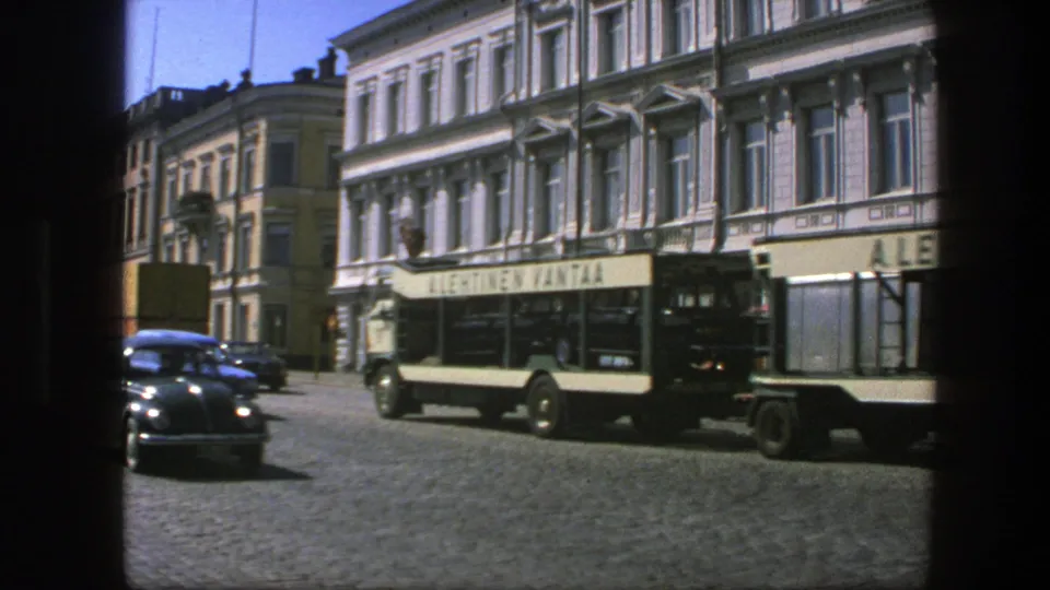 1975 Sweden urban street scene with a tan tour bus, parked cars, storefront awnings, and ornate white buildings seen from a framed viewpoint.