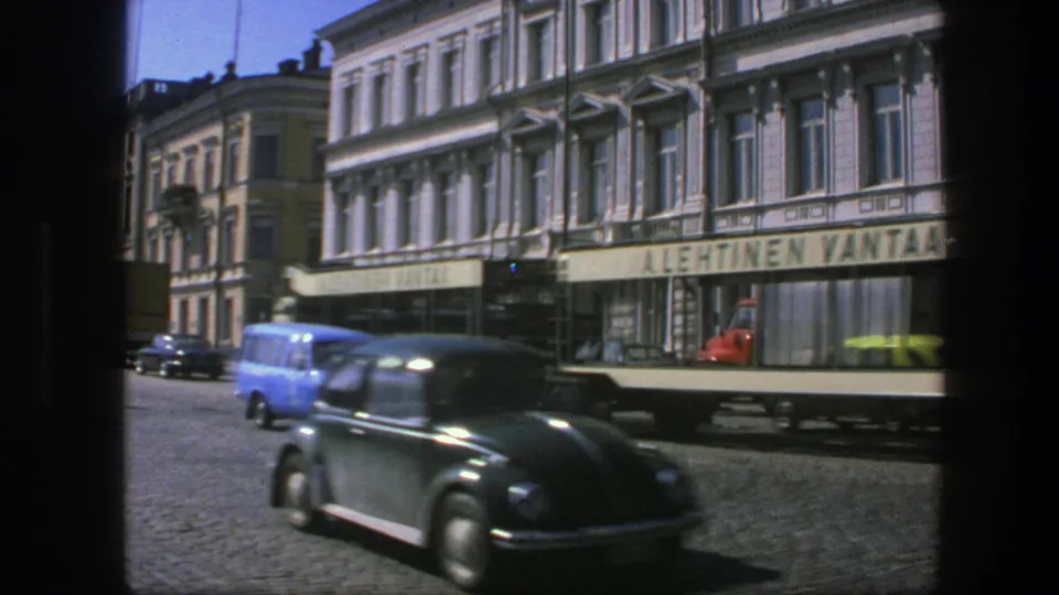 1975 Sweden home-movie frame of a streetcar and dark Volkswagen Beetle moving past storefronts and older multi-story buildings.