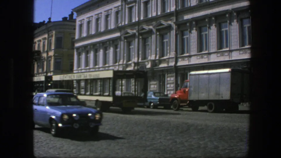1975 Sweden home-movie frame showing a dark sedan near a streetcar, with a red delivery truck and storefronts behind it.