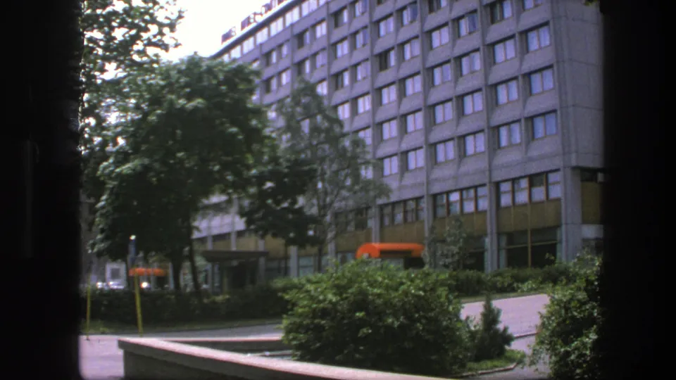 1975 Sweden urban view of a large gridded building facade with trees, shrubs, sidewalk, and orange awnings or vehicles near the entrance.