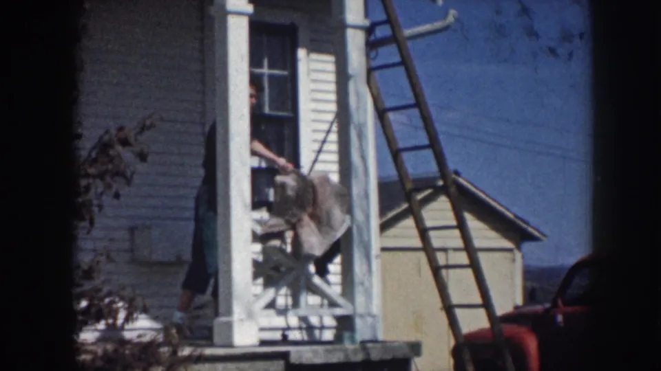 1958 Nebraska home-movie frame showing a woman partly visible on a white clapboard porch near shutters and a leaning ladder.