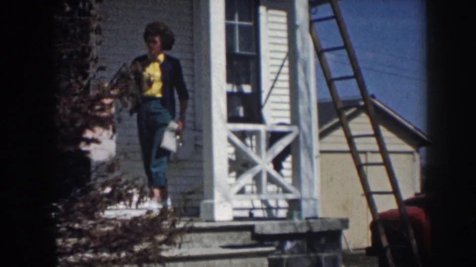 1958 Nebraska frame of a woman in a yellow shirt and dark cardigan on a white house porch, holding a light garment.