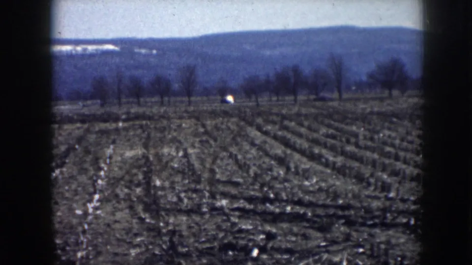 1958 Nebraska rural frame showing plowed field rows, bare trees, a small light structure, and distant hills.