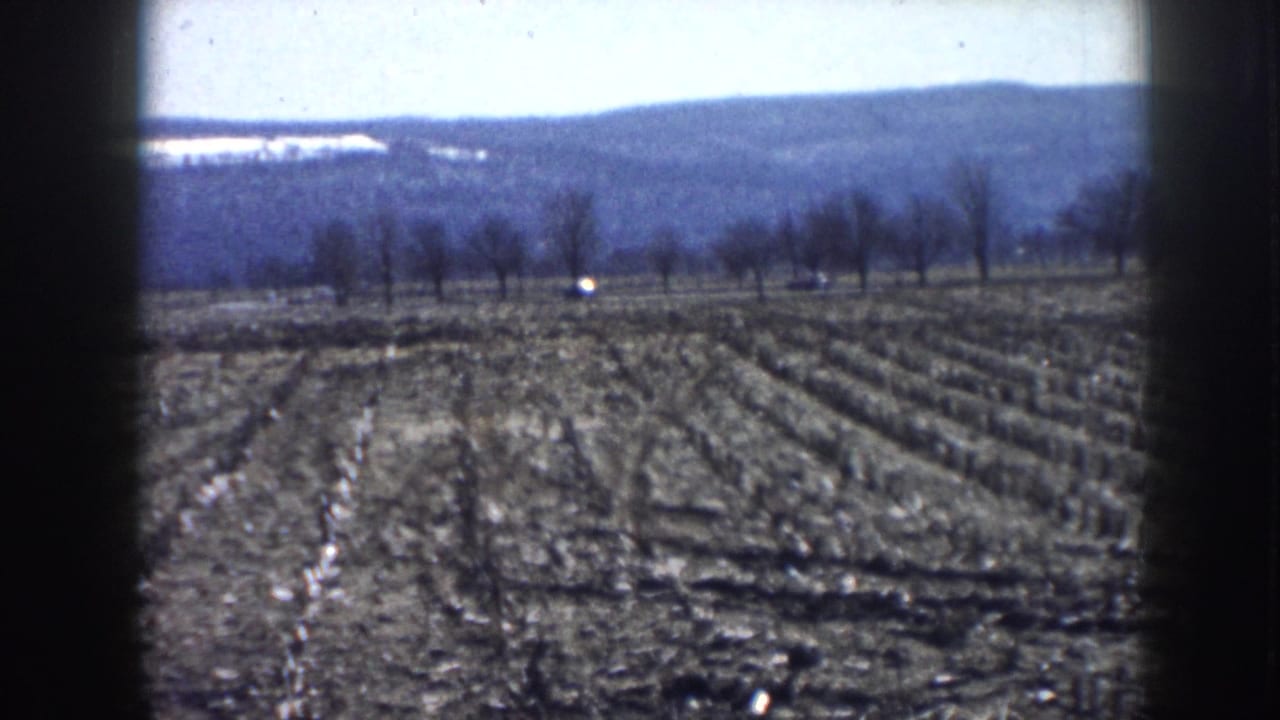 1958 Nebraska home-movie frame showing two cars on a country road past a plowed field, bare trees, and low distant hills.
