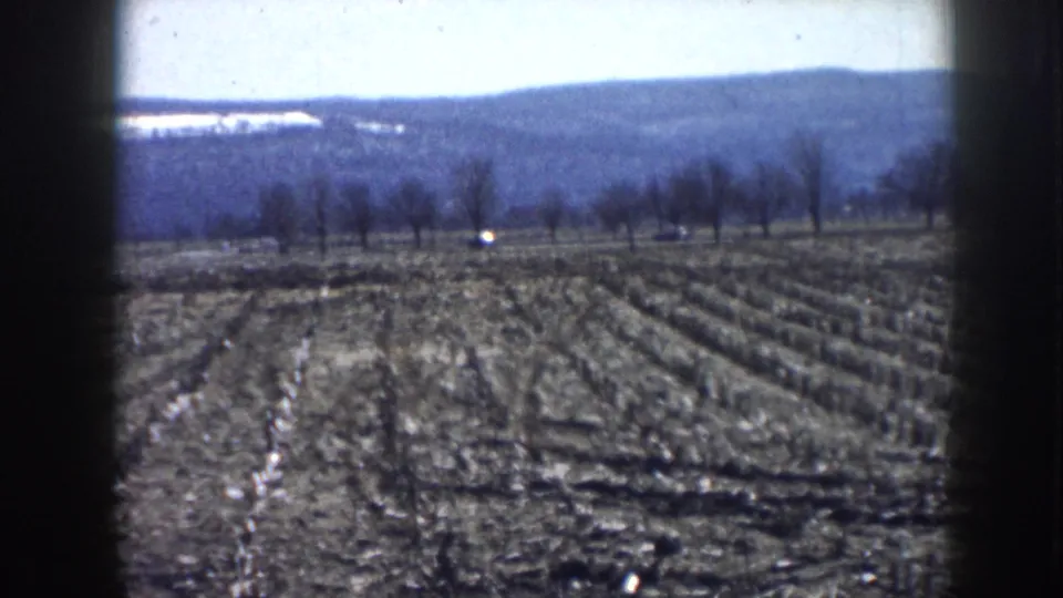 1958 Nebraska home-movie frame showing two cars on a country road past a plowed field, bare trees, and low distant hills.