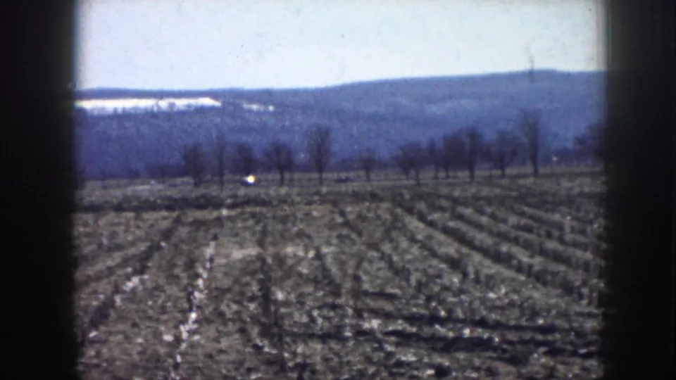 Nebraska rural road scene with cars moving beyond a plowed field, bare trees, and distant hills in soft 8mm color.